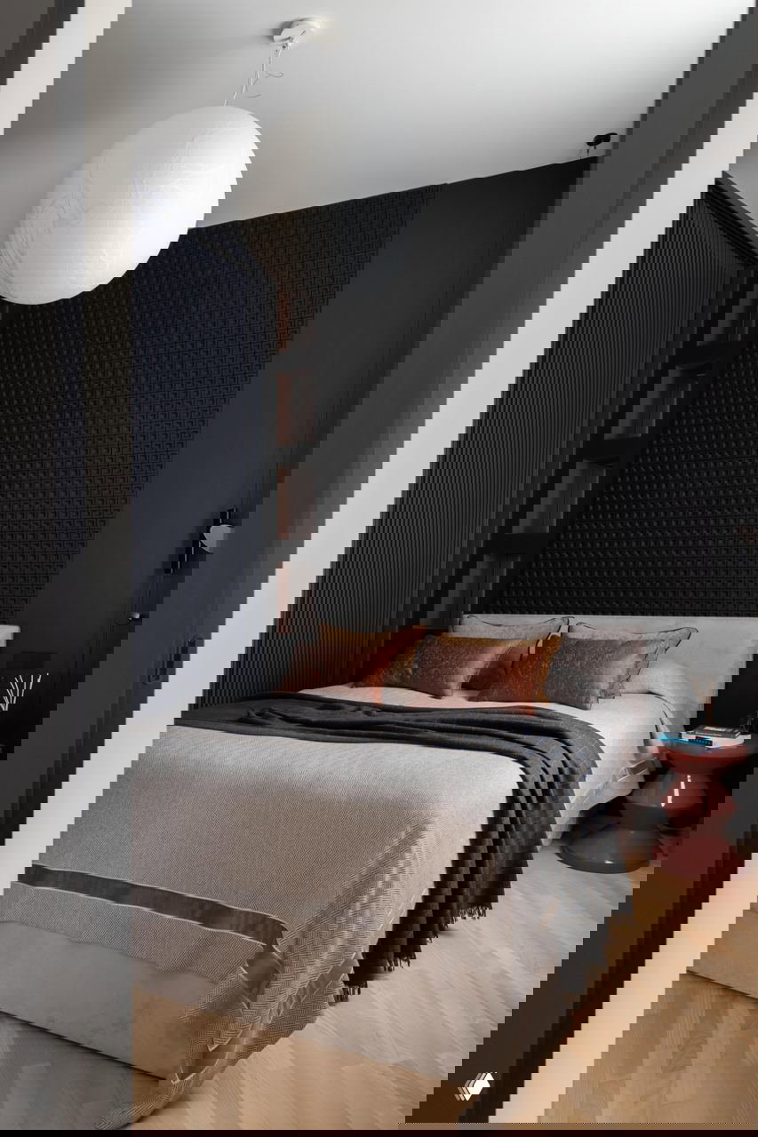 Спальня со сферической подвесной лампой и тёмной текстурной панелью за изголовьем, мягкое покрывало и декоративные подушки создают уютный акцент.