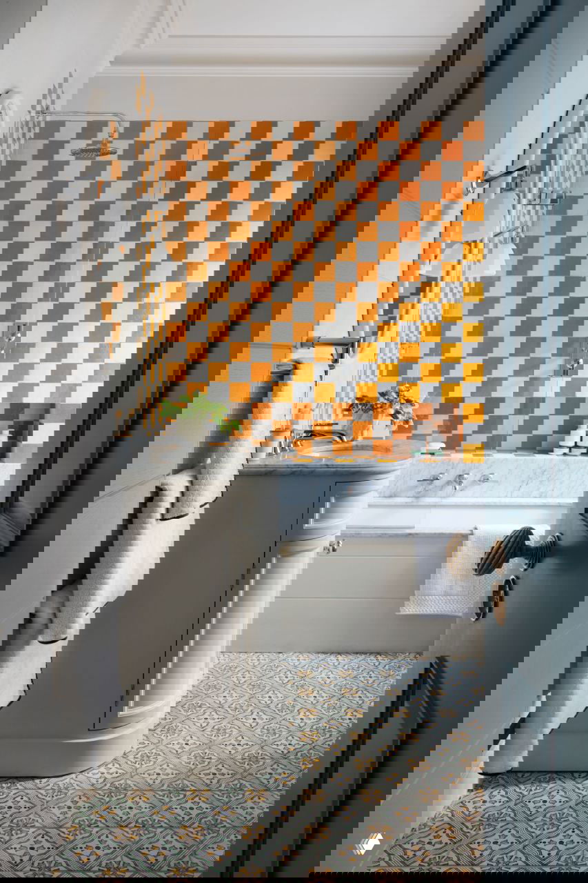 Ванная комната с оранжево‑белой шахматной плиткой в зоне ванны, мраморной столешницей и узорным напольным кафелем — смелое сочетание цвета и узора.