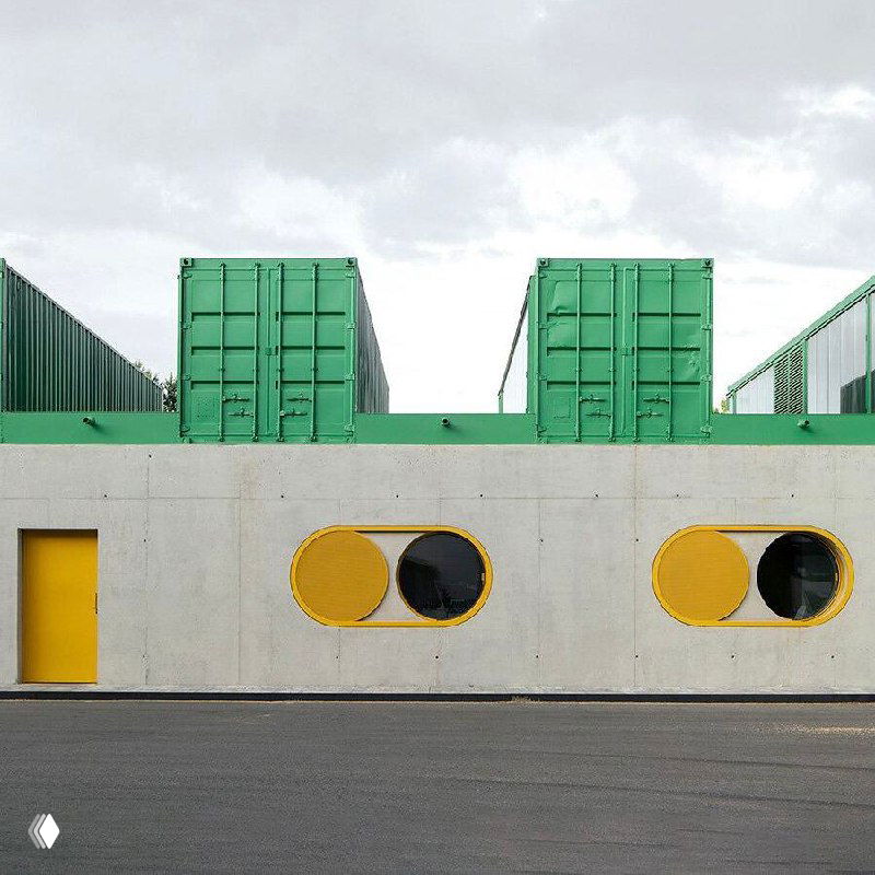 Крупный план бетонной стены офиса с овальными проёмами и жёлтыми рамами на фоне зелёных контейнеров и индустриального неба.