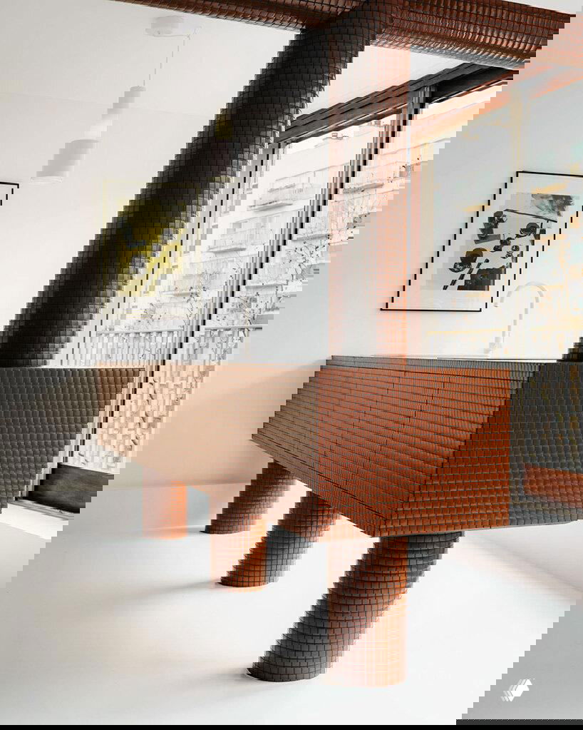 Кухонный или сантехнический модуль, облицованный маленькой коричневой плиткой: массивный стол‑блок на круглых опорах и панорамное окно на заднем плане.