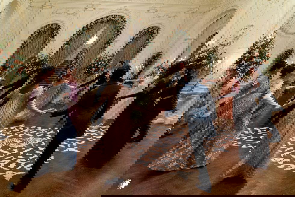 Бал в роскошном интерьере с неоклассическим декором: гости в бальном платье и костюмах танцуют в зале с цветочными композициями и лепниной.