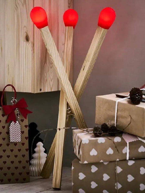 Крупный план дизайнерской лампы в форме трех спичек с ярко-красными пластиковыми головками на фоне нейтральной стены и текстиля в скандинавском стиле.