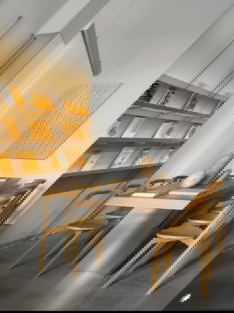 Длинный стол и стулья из светлого дерева у стены с подсветкой полок; минималистичный интерьер, керамические объекты и ровные световые акценты в пространстве.