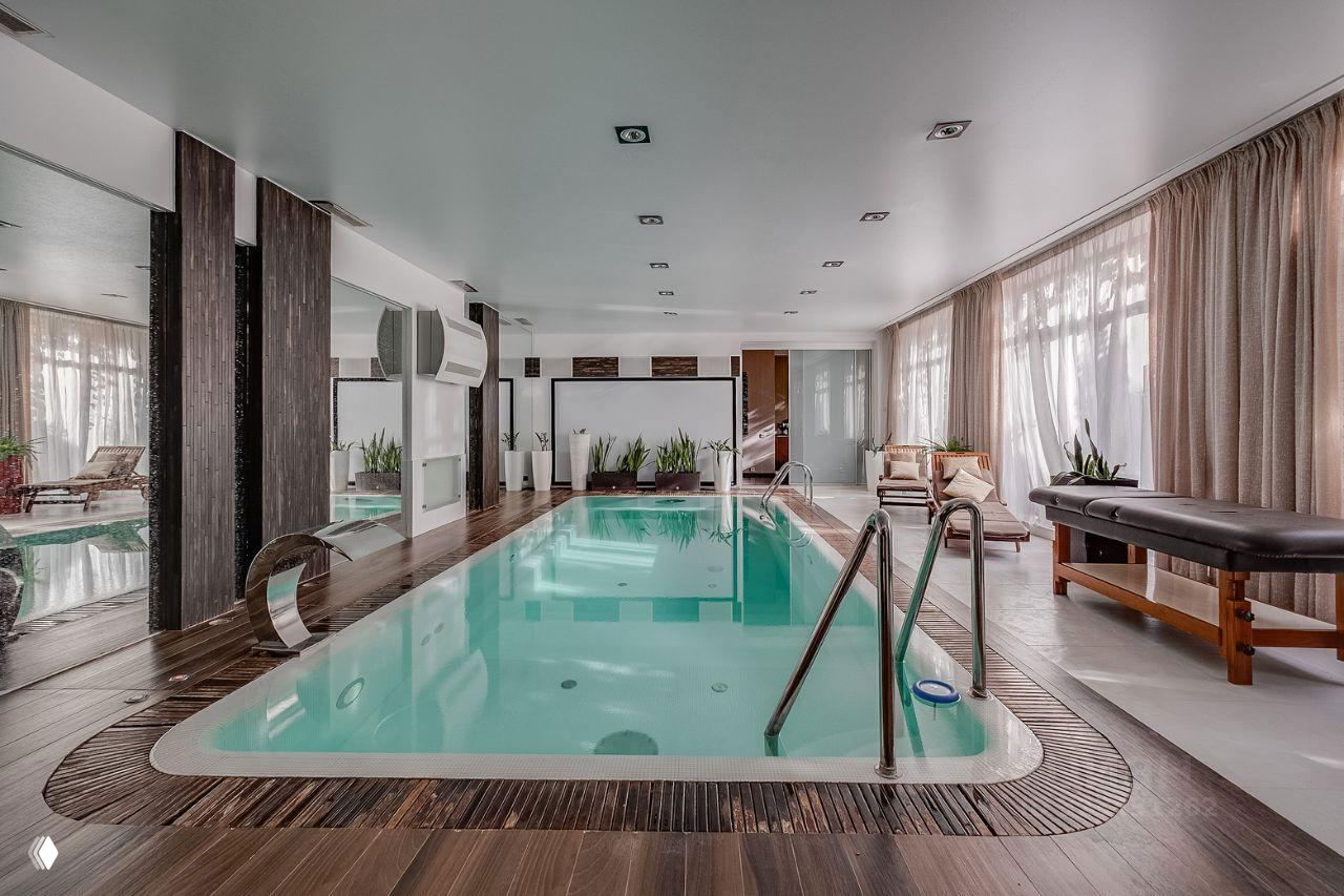 Спортивный зал с крытым бассейном, зеркальными стенами, деревянной отделкой у кромки воды и шезлонгами у панорамных окон для релаксации.