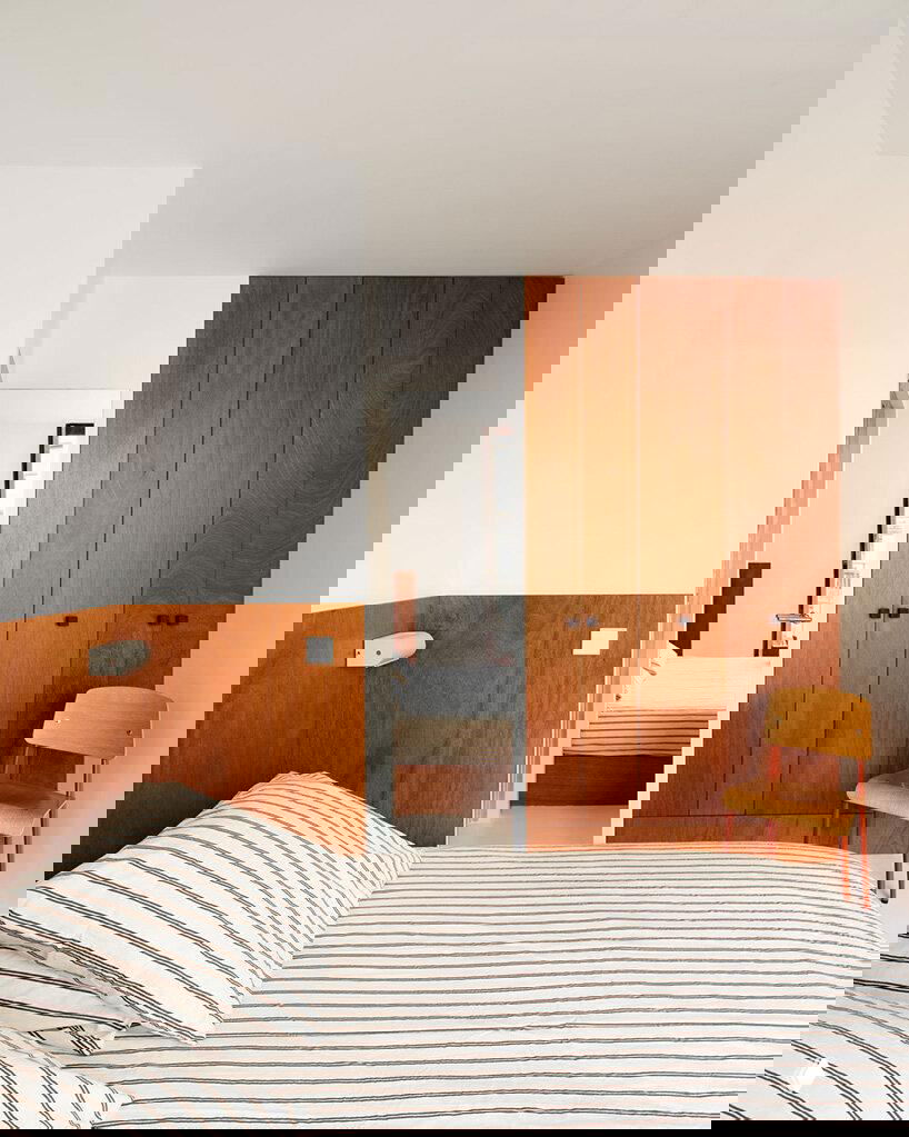 Спальная зона: низкая кровать со светлым полосатым текстилем, встроенный шкаф и деревянные панели из фанеры, минималистичный интерьер в теплых тонах.
