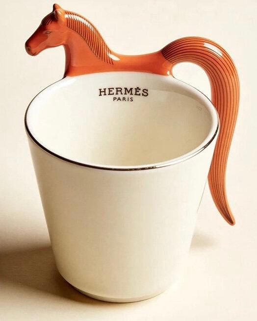 Крупный план керамической чашки из набора Hermès x Bialetti: белая поверхность, внутри логотип Hermès и изящная оранжевая ручка в форме головы и шеи лошади.