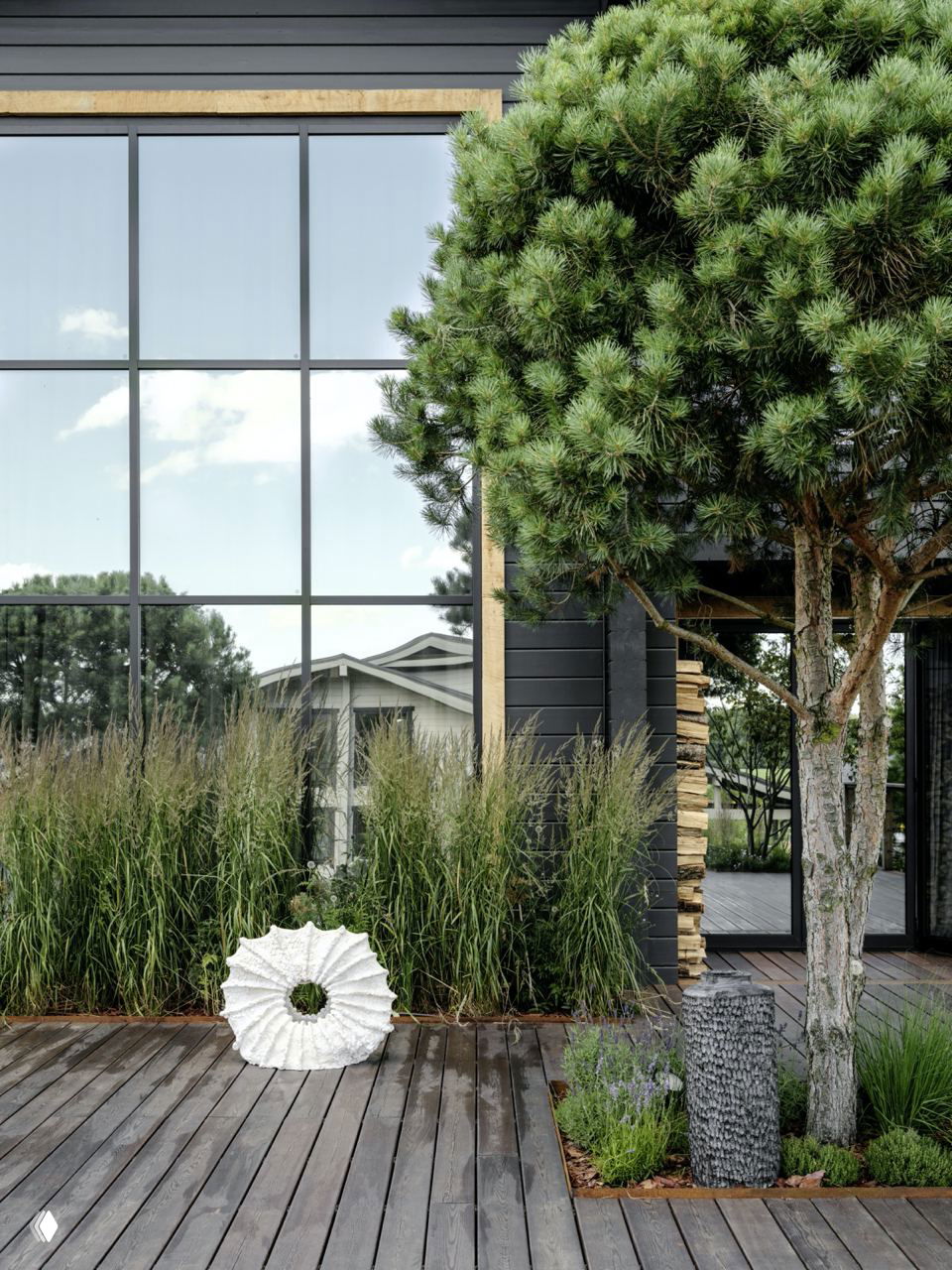Экстерьерная зона с большой зеркальной оконной панелью, деревянной террасой и зелёными насаждениями — гармоничное соседство природы и архитектуры.