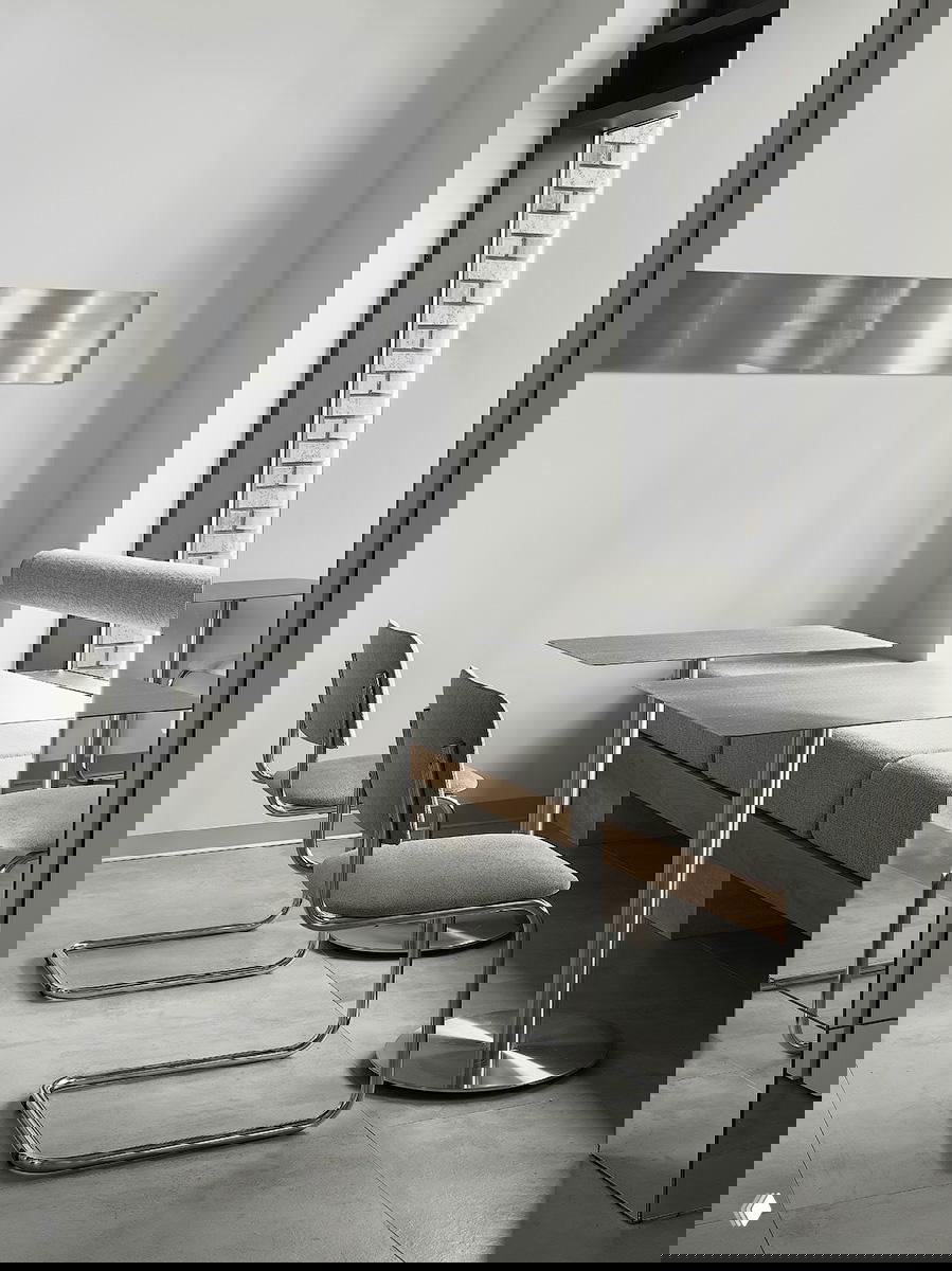 Уютный уголок с серыми сиденьями и металлическими столиками у окна: чистые линии, нейтральная палитра и отражающая металлическая полоса на стене, усиливающая ритм света.