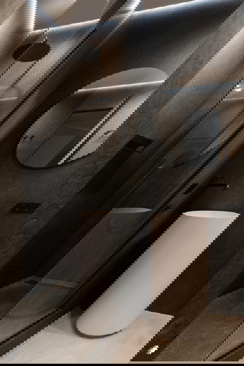 Тёмная ванная комната с фактурной отделкой, стеклянной душевой кабинкой, круглым зеркалом и белой цилиндрической раковиной на контрастном фоне.