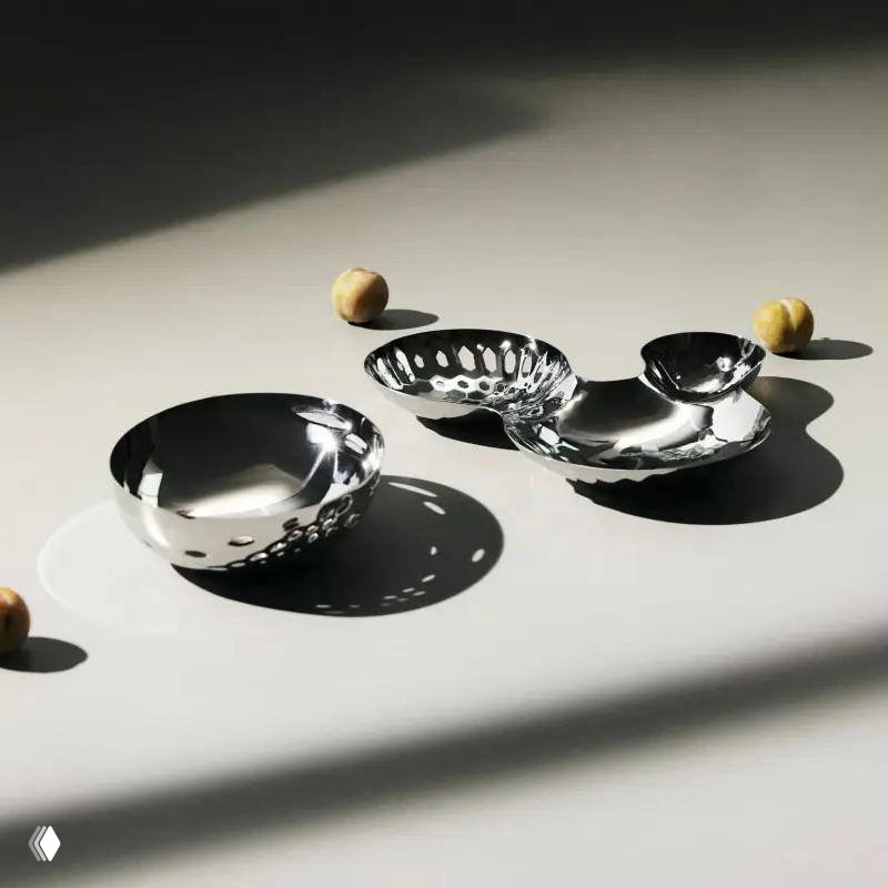 Несколько лаконичных стальных чаш и блюдец на световом фоне, игра света и тени на поверхности минималистичной коллекции посуды для сервировки.