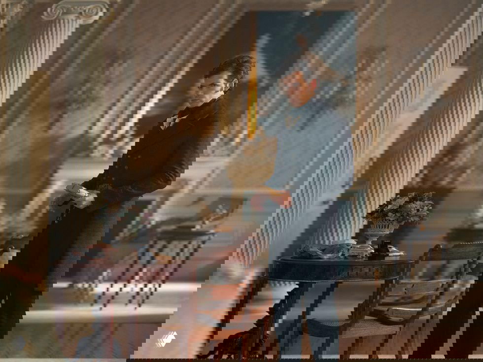 Интерьер с персонажем в кадре: антикварная мебель, столы и декоративные элементы в неоклассическом стиле, атмосферный кинематографический свет.