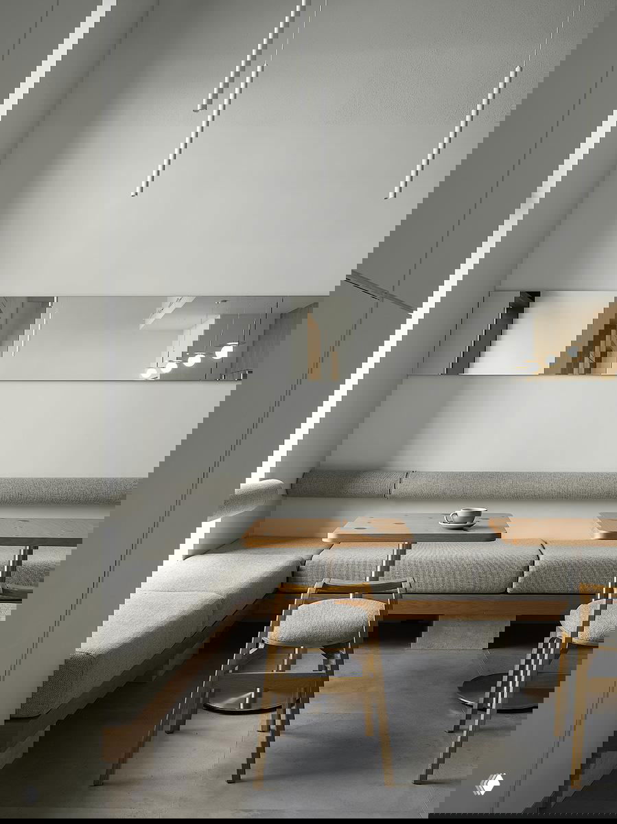 Светлый зал с высокими потолками, продолговатыми подвесными светильниками и зеркальной панелью, создающими визуальную глубину и спокойный интерьерный ритм.