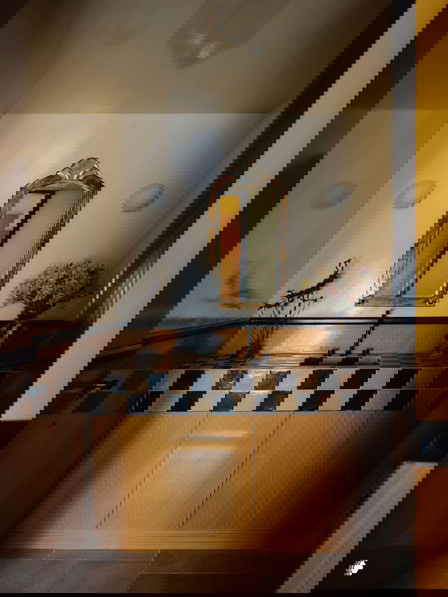 Деталь интерьера: умывальник с зеркалом и плиткой в шахматный узор, ваза с цветами и деревянная отделка стен в ванной зоне заведения.