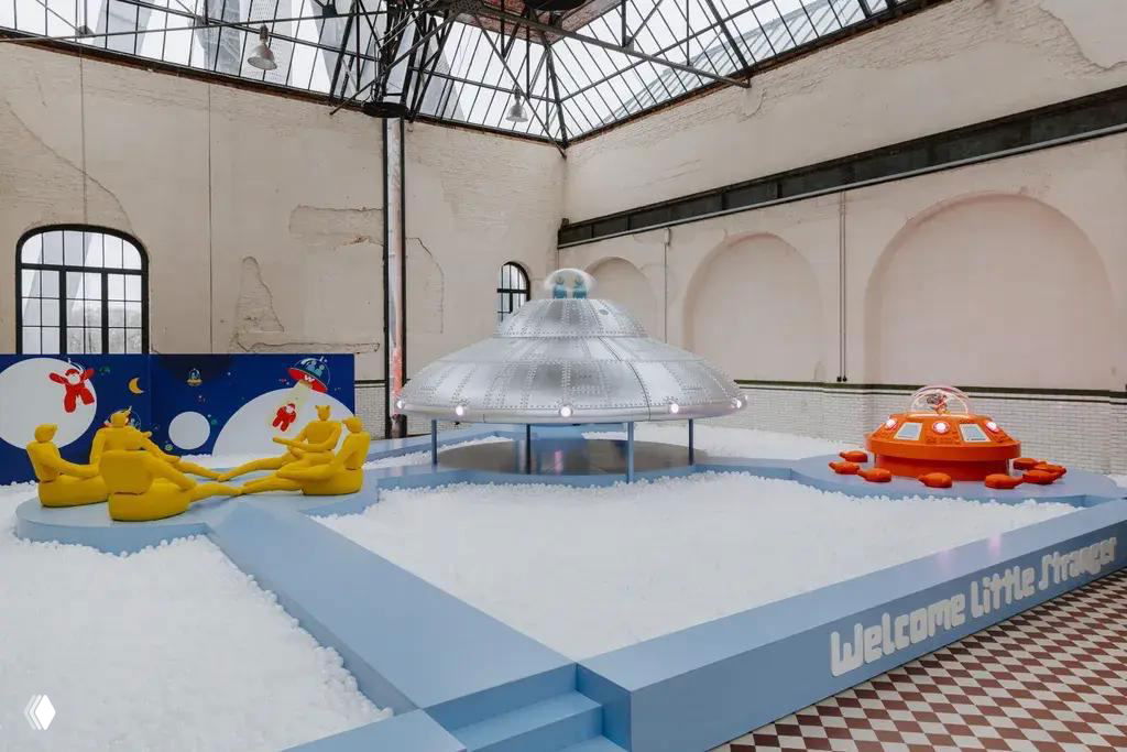 Общий вид игровой экспозиции в зале бывшей шахты: серебристая тарелка, оранжевый игровой модуль, желтые сиденья и бассейн из белых пластиковых шариков под стеклянной крышей.