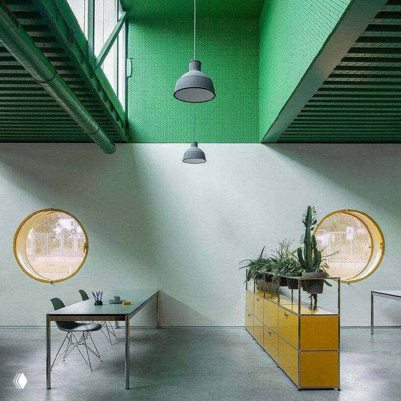 Рабочее пространство с низкими столами, овальными окнами и зелёной структурой крыши: функциональный минимализм и яркие акценты цвета.