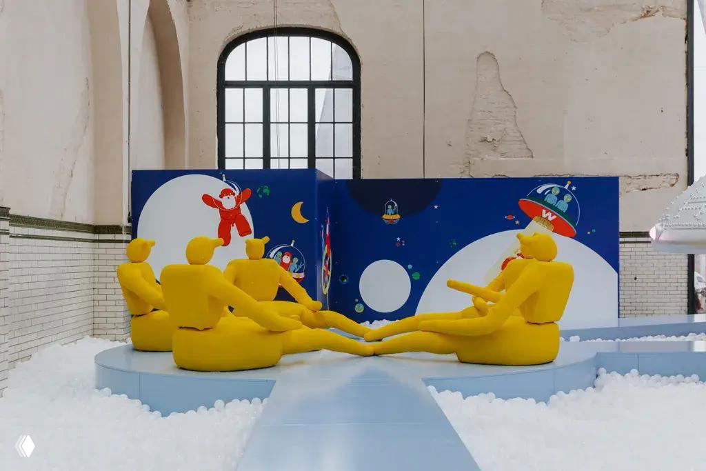 Группа желтых мягких скульптур‑фигур, похожих на отдыхающих пришельцев, расположенных на платформе на фоне декоративной космической графики на стене выставки.