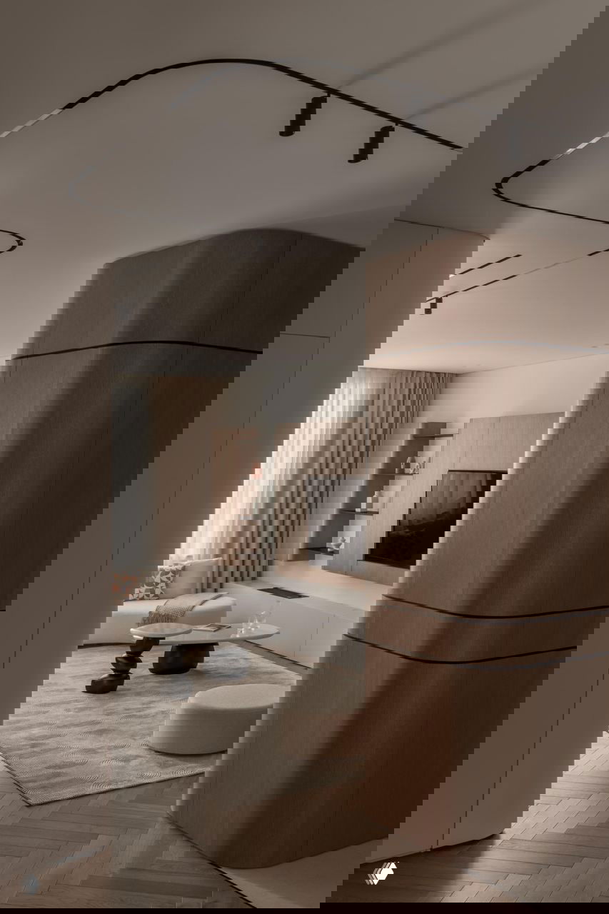 Вид на гостиную с округлённым деревянным пилоном, трековым освещением на потолке, мягкой зоной и кофейными столиками — спокойная тёплая палитра интерьера.