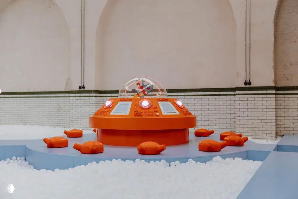 Оранжевый круглый игровой модуль с прозрачным куполом и окружением в виде маленьких подушек, установленный в зоне для детей посреди белого поля шариков.