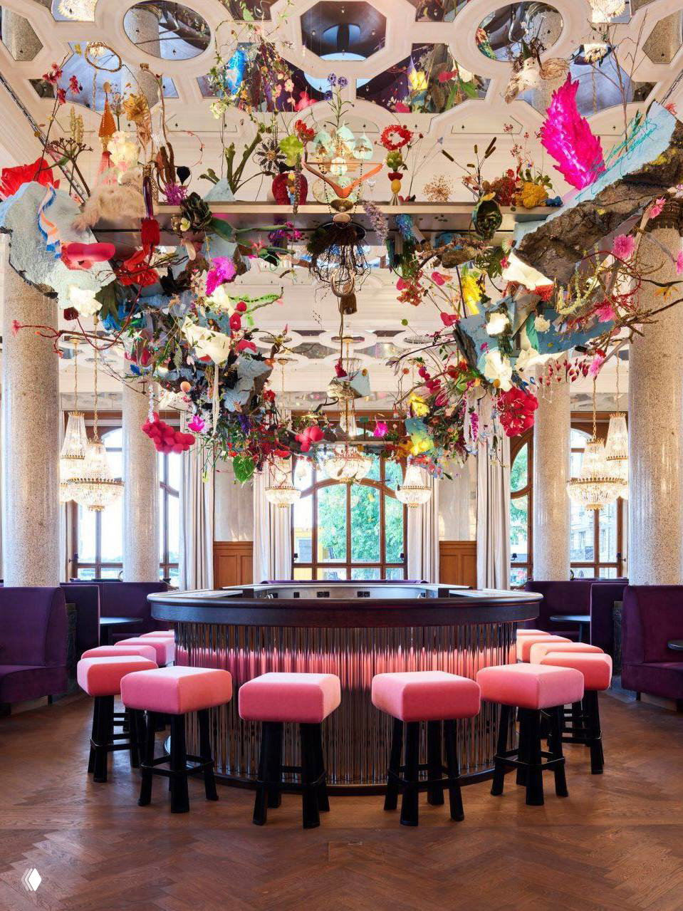 Интерьер нового крыла Les Trois Rois: центральный бар с розовыми табуретами, широким деревянным паркетом, зеркальными вставками и крупной подвесной цветочной инсталляцией.