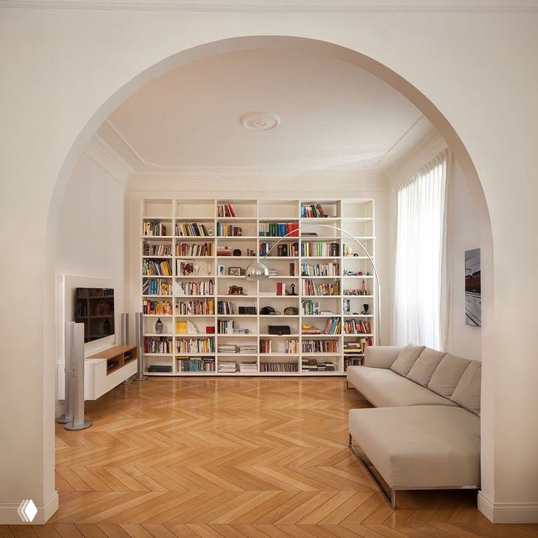Уютная белая гостиная с большой библиотекой в нише, низким диваном и классическим деревянным паркетом, сочетание классики и минимализма.
