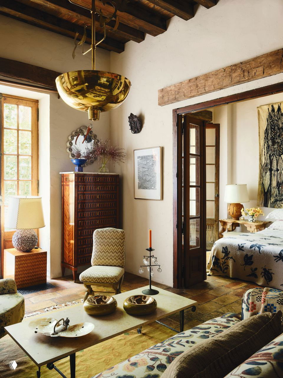 Латунная люстра с декоративными петухами над зоной отдыха: кресло, комод с интарсией, настольная лампа и высокий деревянный потолок.