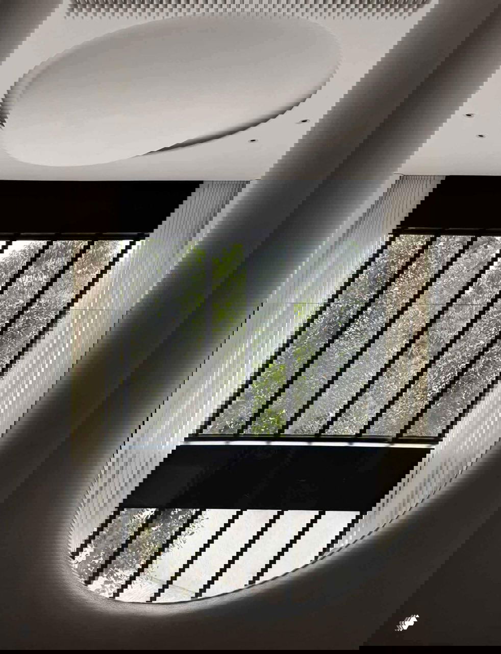 Овальное окно с видом на зелень за стеклом, рамки тёмного металла контрастируют с округлыми формами интерьера и светлыми шторами.