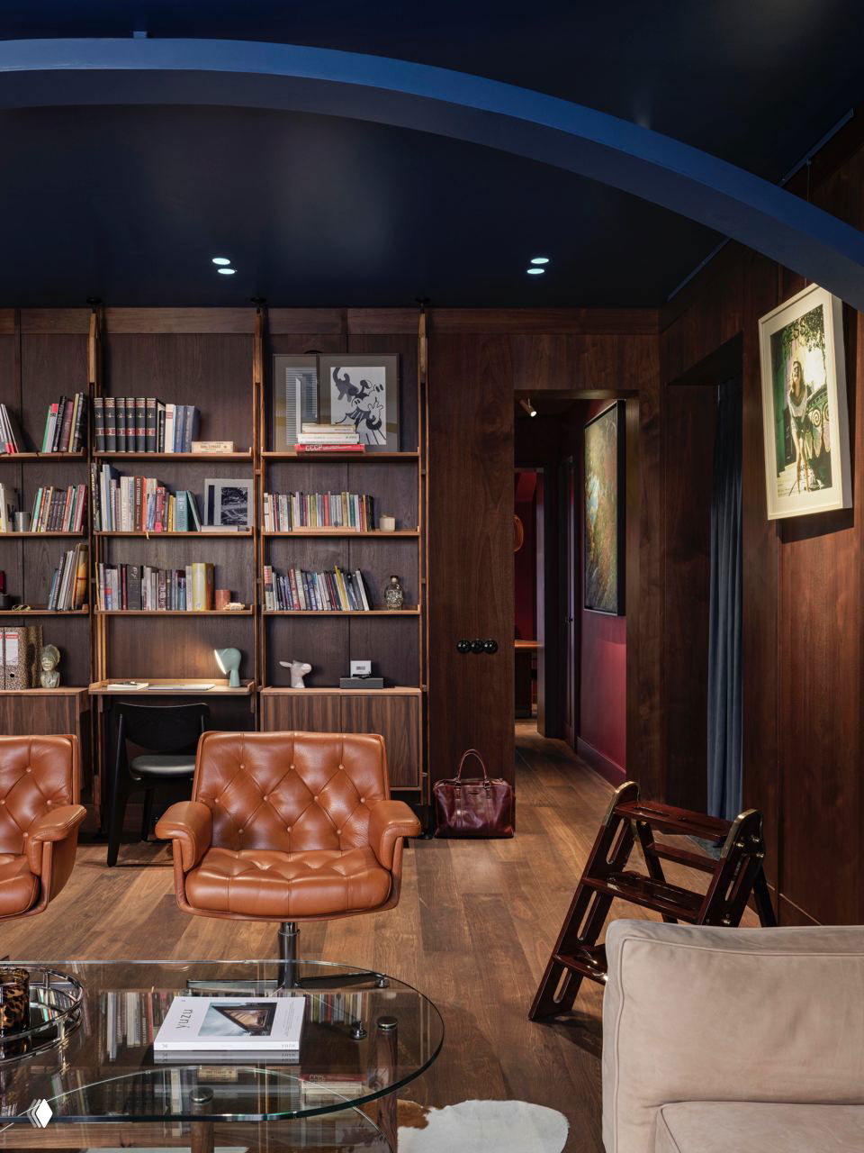 Тёмная библиотека в середине столетия: кожаные стёганые кресла, деревянные стеллажи с книгами, стеклянный журнальный стол и тёплый деревянный пол.