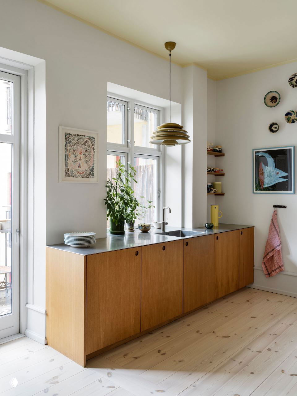 Кухня с низким деревянным кухонным модулем и металлической столешницей, большие окна и минималистичная расстановка предметов на фоне светлых стен и деревянного пола.