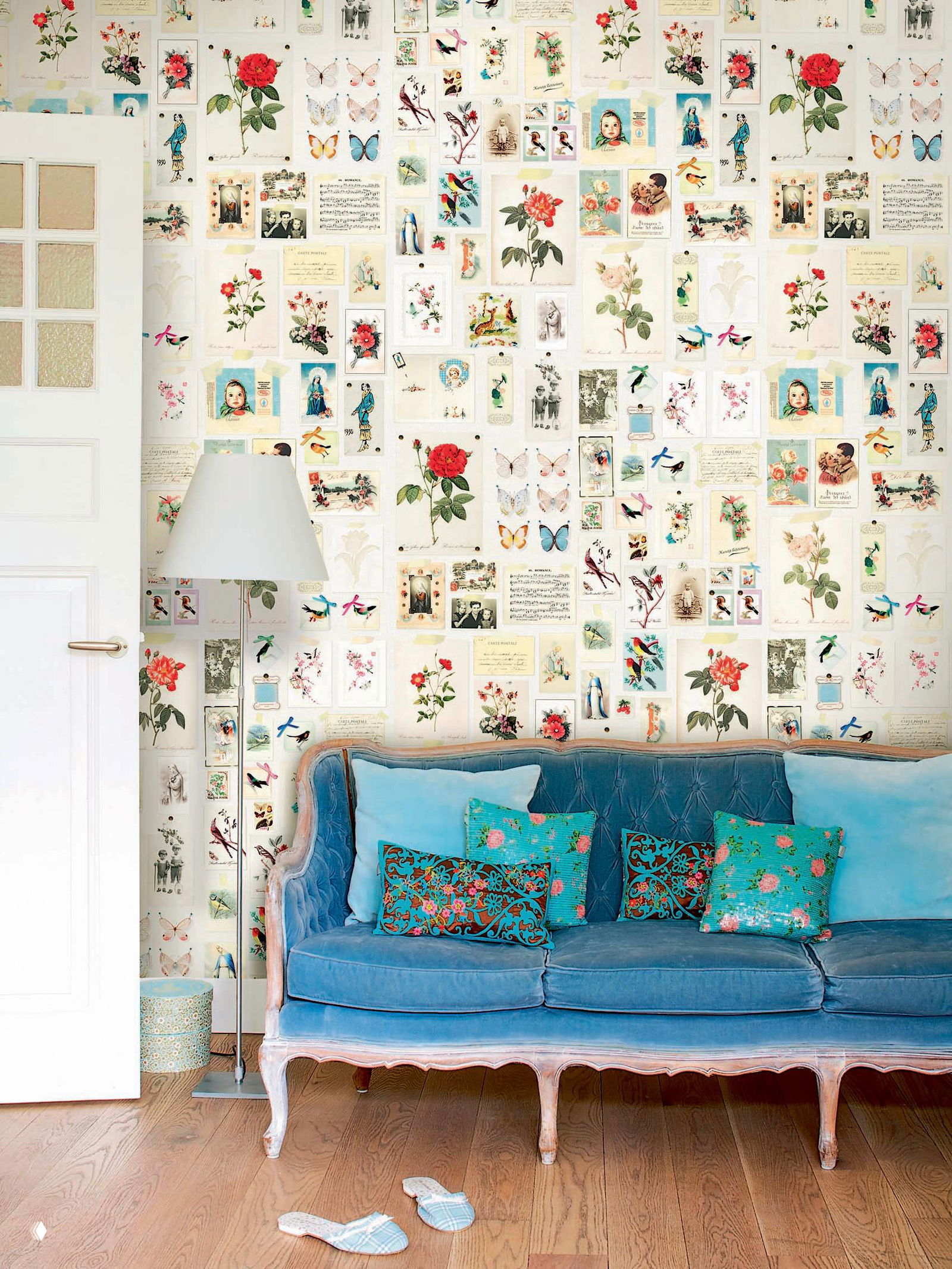 Интерьер с винтажным диваном на фоне обоев, составленных из ретро-иллюстраций и карточек, яркие подушки и лампа дополняют композицию.