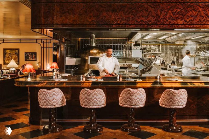 Открытая кухня Gloria Osteria: повар у мраморной стойки, вид на рабочую зону и печь, тёплая атмосферная подсветка и деревянные текстуры.