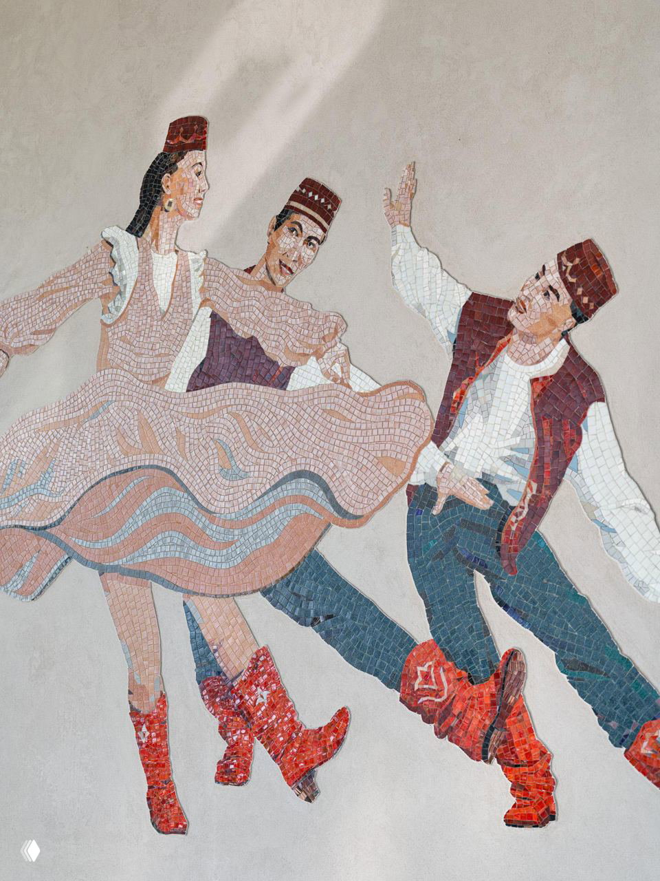 Крупный план мозаичной композиции: фрагмент танцующих людей в нарядах, выполненных из мелких цветных плиток, перед ней светлые табуреты.