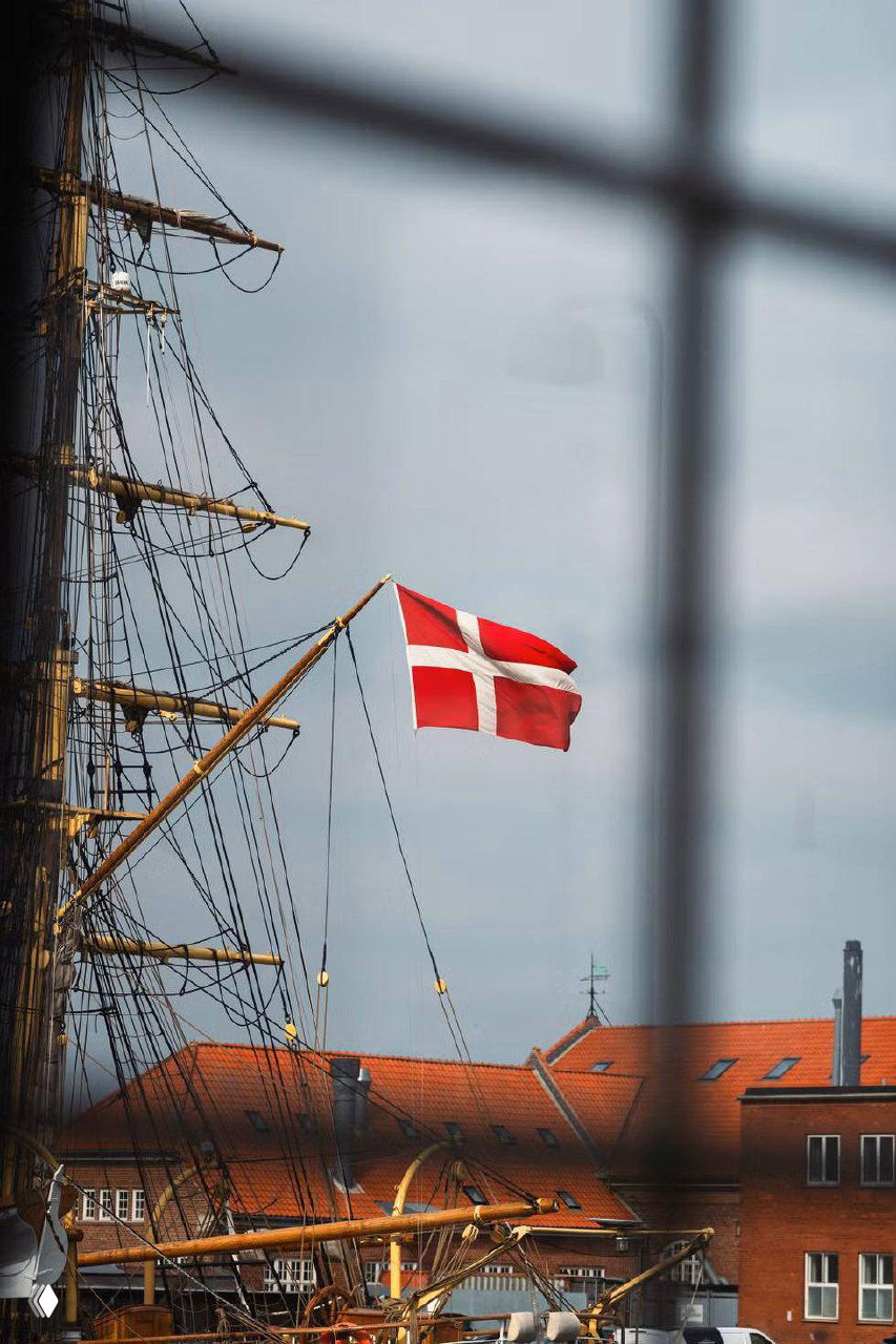 Корабельные мачты и флаги на фоне городской застройки, фотография содержит цифровые артефакты и искажения.