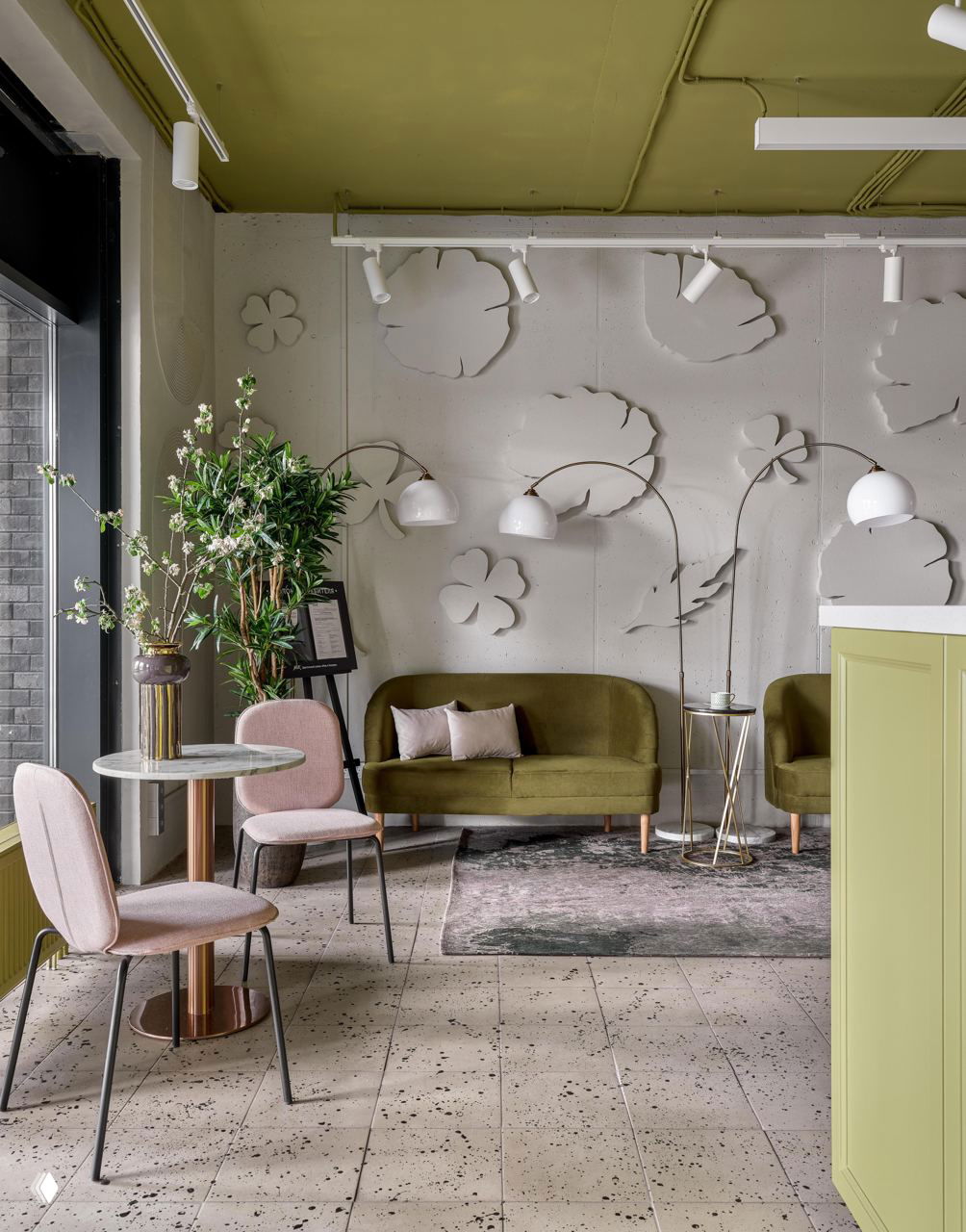 Зона отдыха в бутике: оливковый диван, розовые стулья, рельефные декоративные элементы на светлой стене и зелёные акценты.