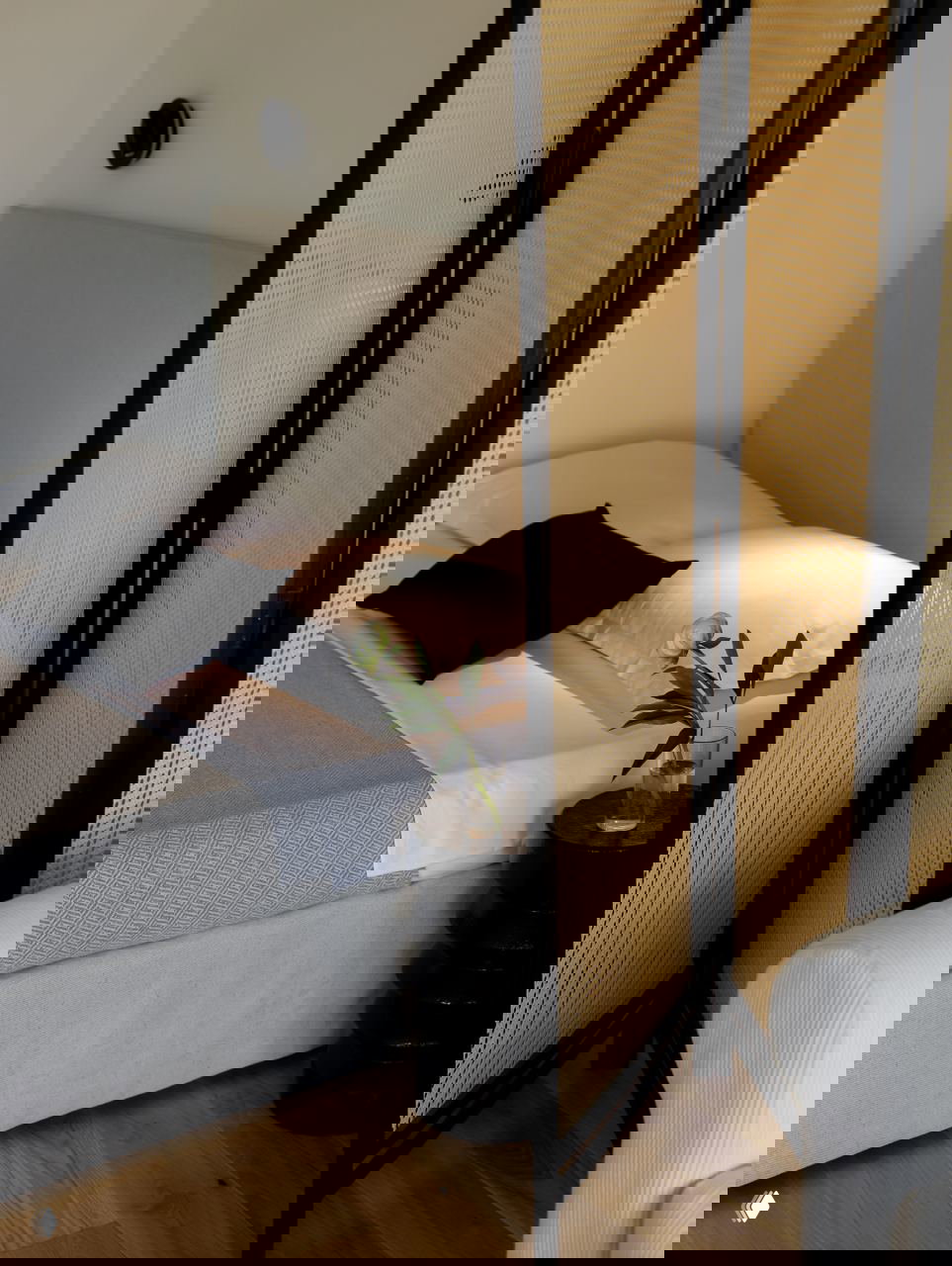 Крупный план прикроватной зоны: мягкая постель, декоративная чёрная подушка и маленький чёрный столик с вазой и цветком рядом со ширмой.