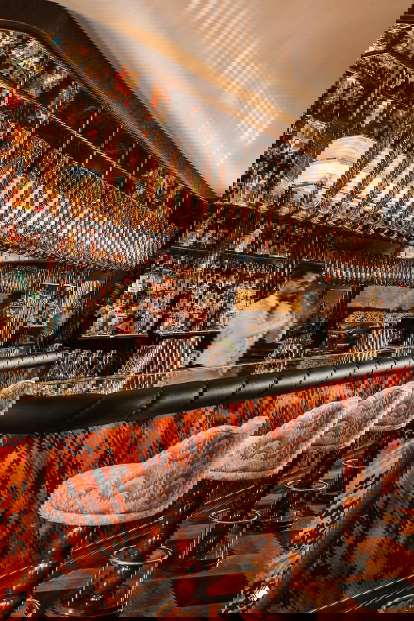 Барная зона ресторана: длинная стойка, мягкие высокие табуреты с узорной обивкой и подвесные стеклянные светильники над баром.