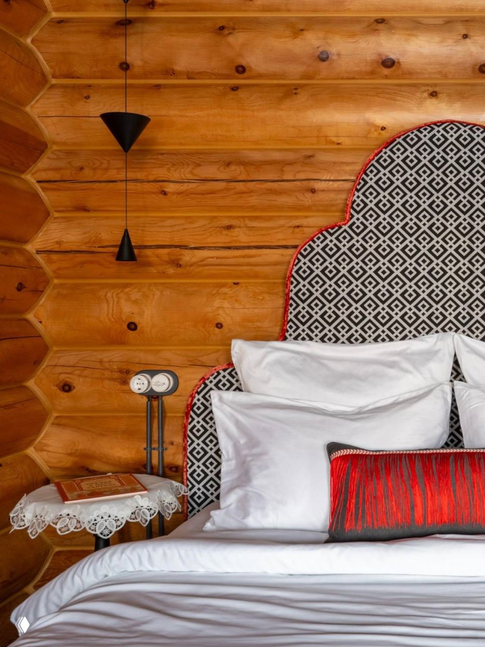Изголовье кровати с орнаментальной обивкой на фоне кедровых бревен, белые подушки и декоративная подушка с красной бахромой — гармония узора и текстуры