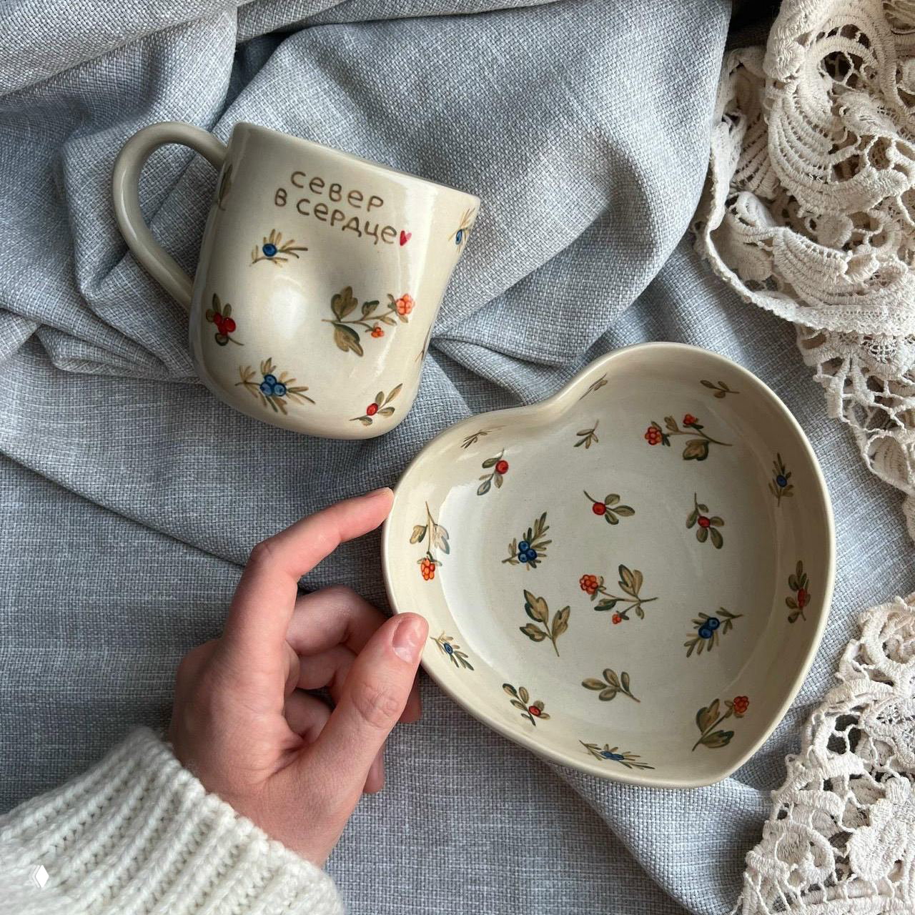 Ручная керамическая кружка с надписью «север в сердце» и тарелка в форме сердца с мелким растительным орнаментом на светлом текстильном фоне.