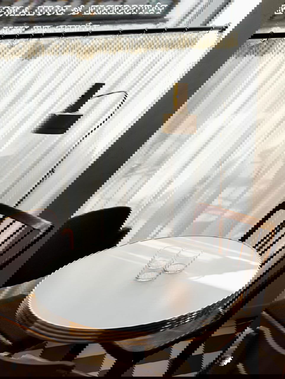 Круглый белый столик у окна с двумя стаканами и настольной лампой, лёгкая занавеска создаёт уютную атмосферу у окна кафе.