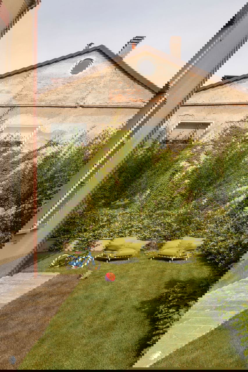 Зелёный частный сад миланской квартиры с низкими зелёными креслами, детским велосипедом и газоном на фоне старинного фасада дома.