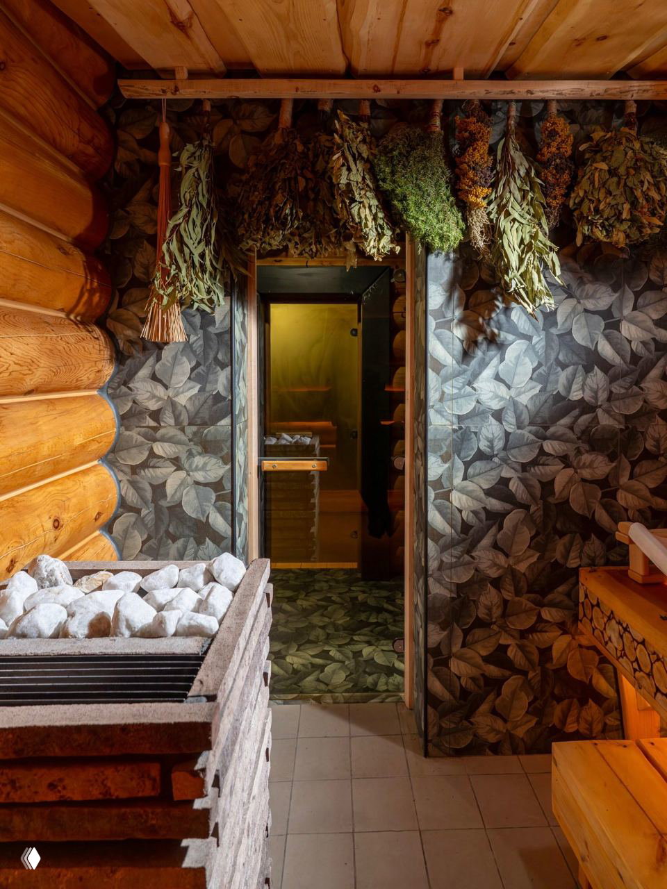 Интерьер парной и предбанника: висящие веники, каменка на первом плане и декоративные обои с крупными листовыми мотивами в переходной зоне