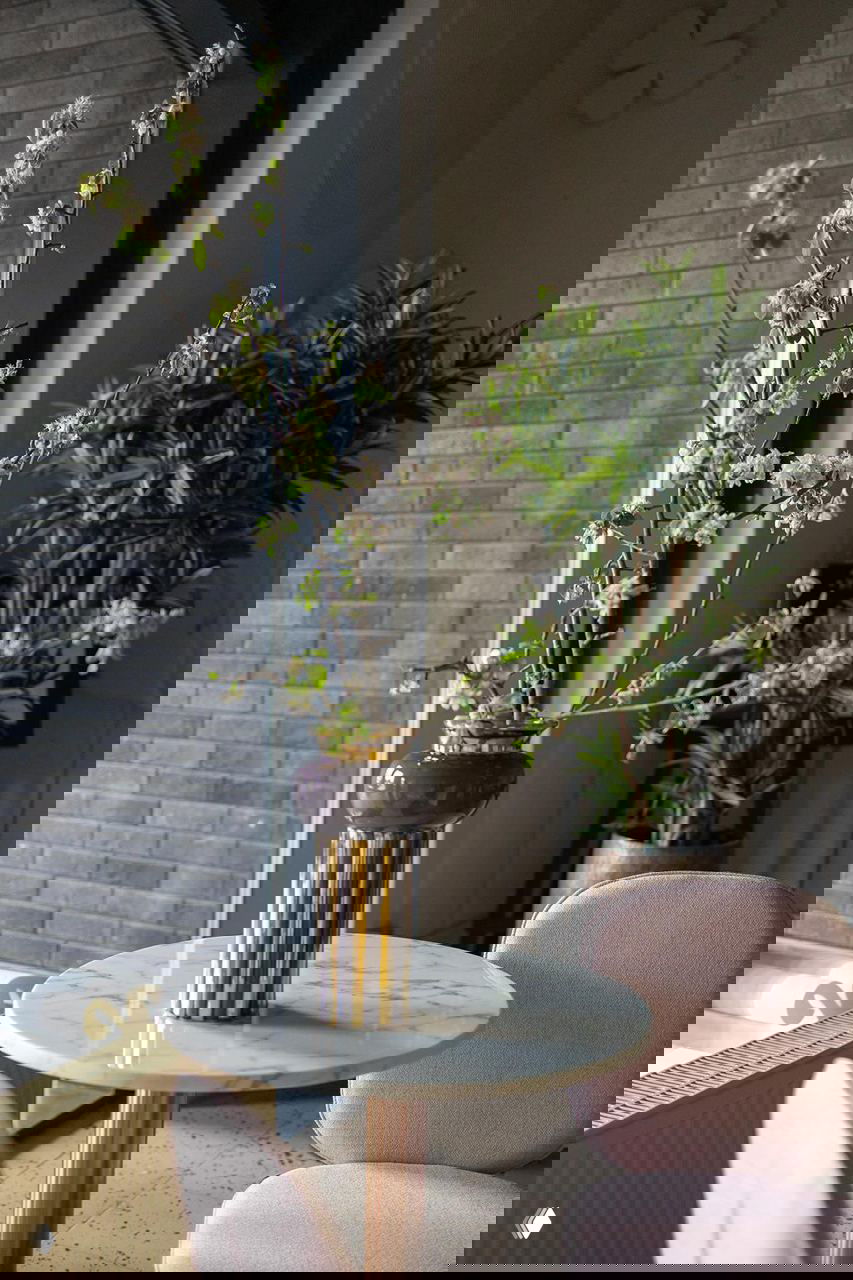 Небольшой столик у окна с вазой и весенними ветками, на заднем плане зелёные растения у панорамного окна бутика.