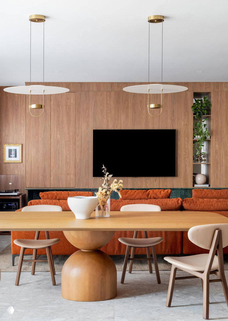 Обеденная зона на фоне деревянной облицовки: круглый деревянный стол, стулья и терракотовый диван с подвесными светильниками над столом.