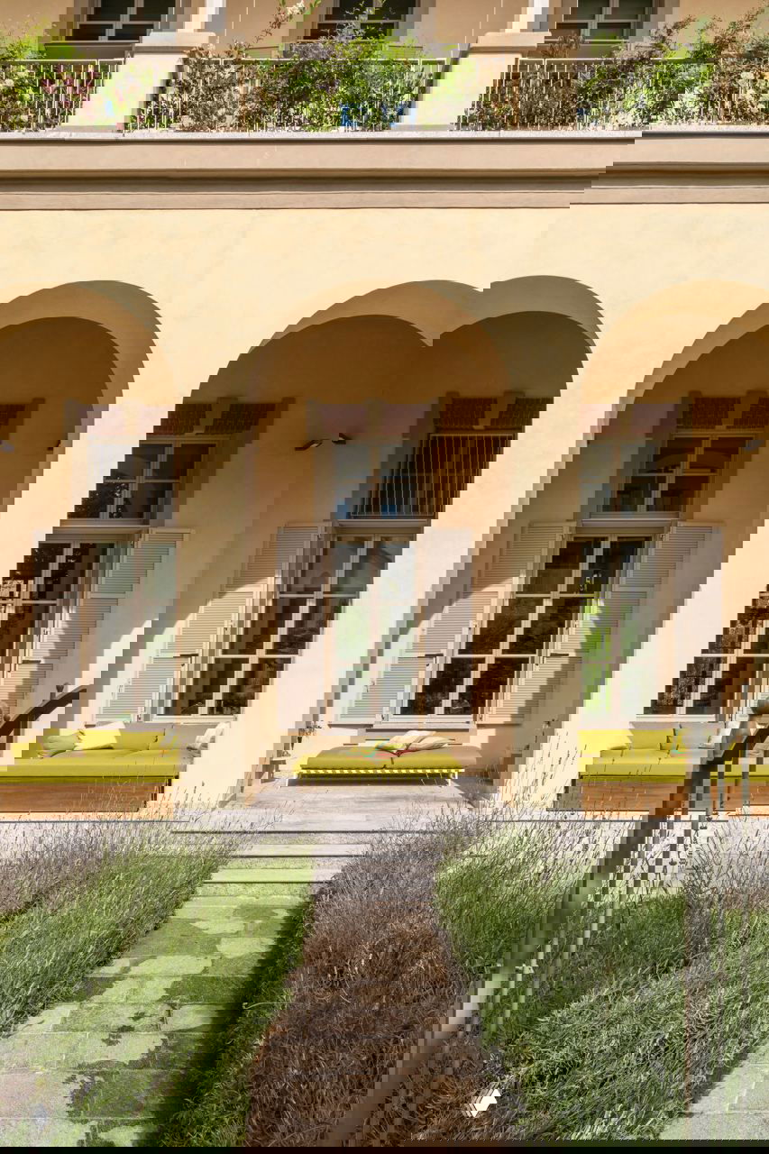 Фасад дома с арочными окнами и лавками с оливковой обивкой на веранде; симметрия и мягкие природные оттенки в экстерьере.