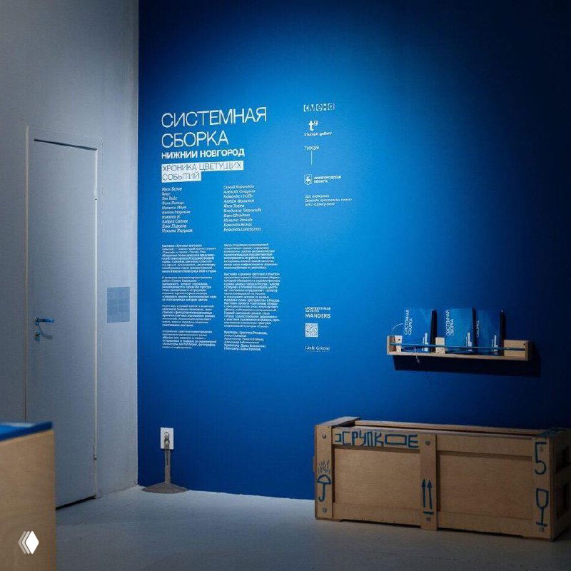 Голубая стеновая панель экспозиции с заголовком «СИСТЕМНАЯ СБОРКА», рядом деревянные ящики для перевозки арт‑объектов и стойка с буклетами выставки