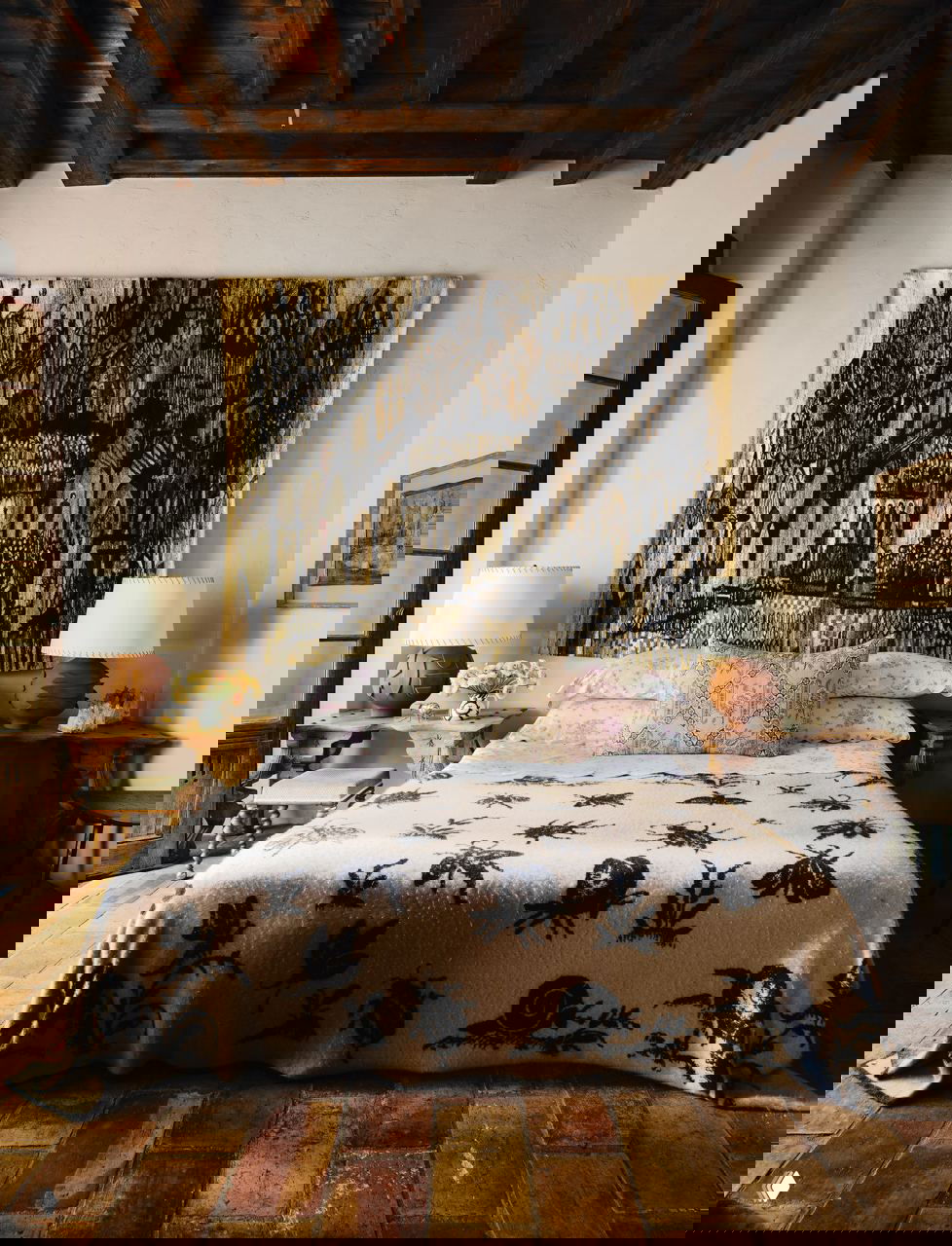 Спальня в прованс-стиле: кровать с растительным покрывалом, декоративные подушки и большое тканевое панно на оштукатуренной стене под деревянными балками.