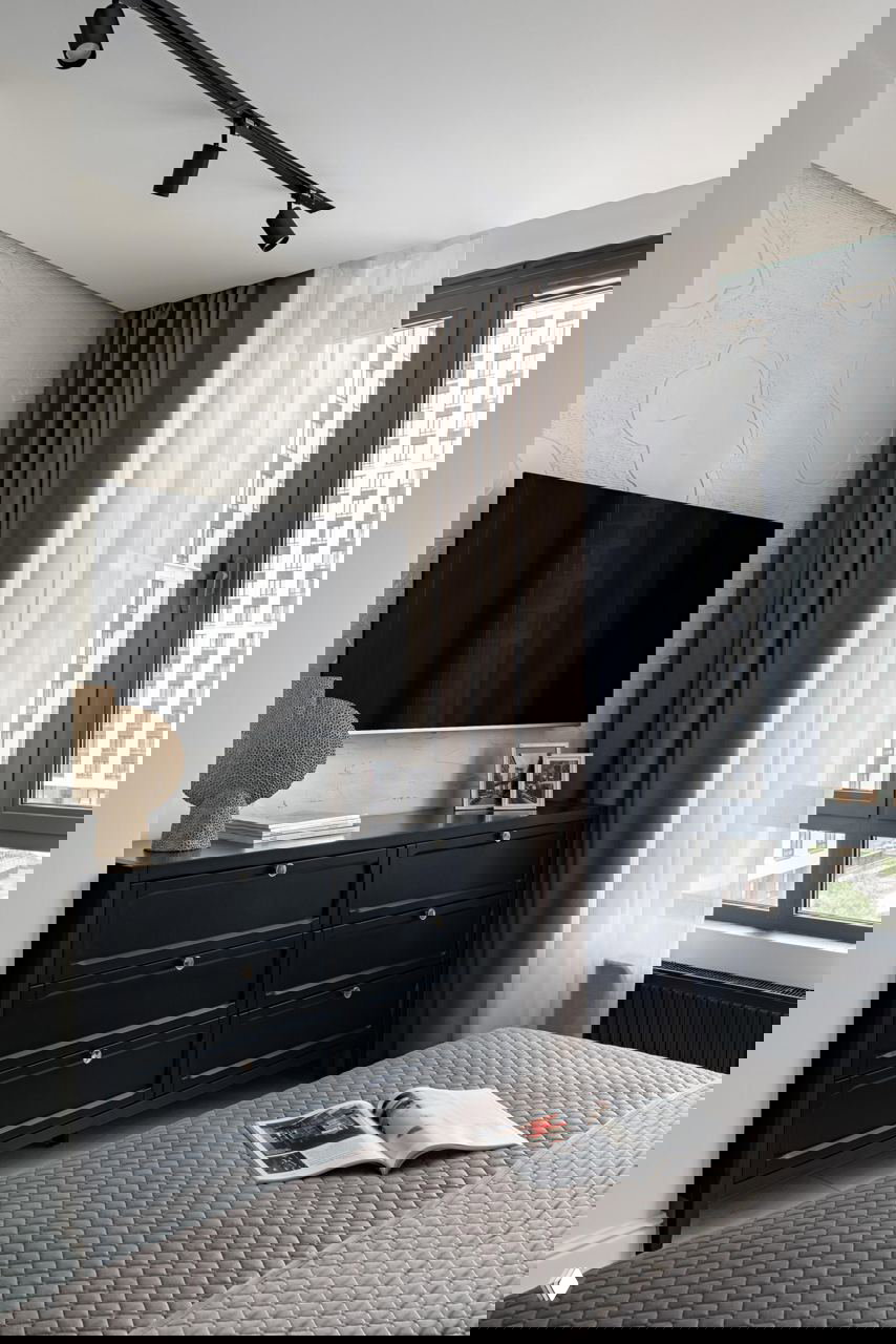 Спальня с тёмным комодом под настенным телевизором, рельефная отделка стены с морским мотивом и аккуратное постельное покрытие при дневном свете.