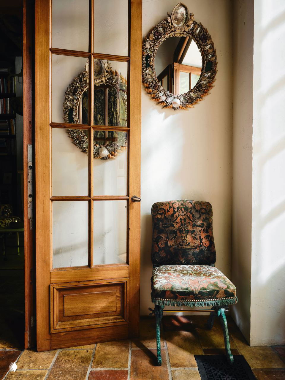 Деревянная дверь со стеклянными створками, круглое зеркало в раме из ракушек и винтажный стул у солнечного оконного проема в комнате.