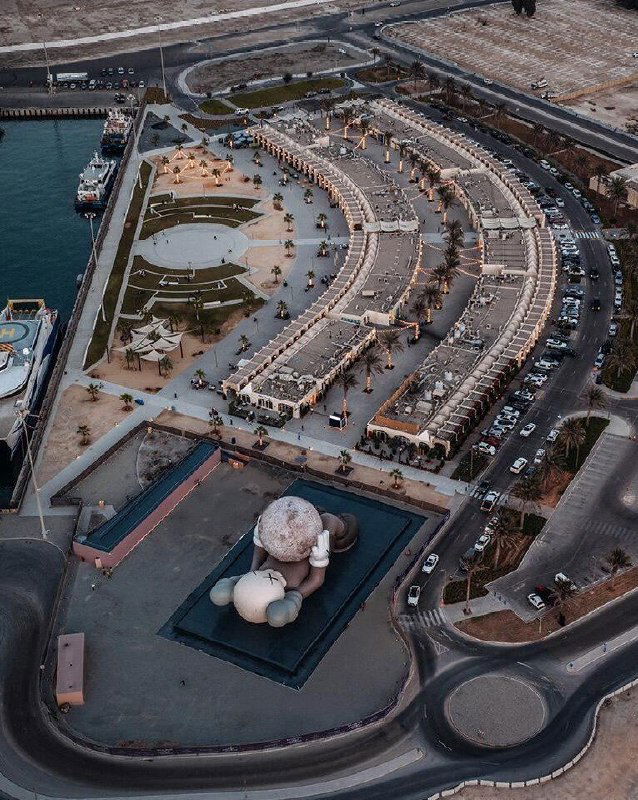 Аэрофотоснимок инсталляции KAWS в порту Мина Зайед: лежащая фигура длиной 32 метра на платформе у воды, окружающая инфраструктура и парковки.