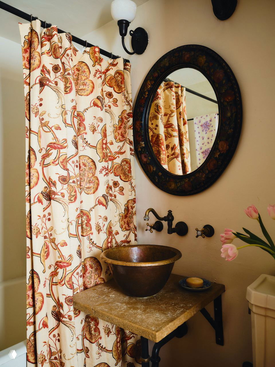 Ванная зона с занавеской в цветочный узор, круглым зеркалом и бронзовой раковиной на каменной столешнице — компактное оформление в прованс-духе.