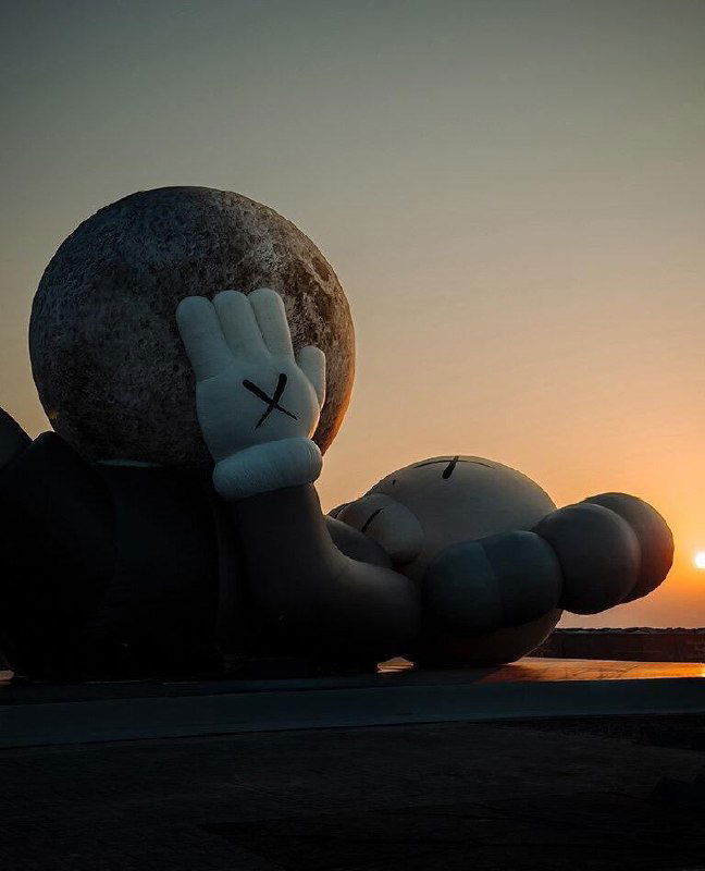 Крупный план головы и руки персонажа KAWS с символической «луной» в ладони на фоне заката над заливом, теплые тональные градации.